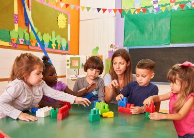 Kindergarten teacher and children play with building blocks together in daycare or preschool